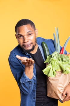 African american male model gives air kisses against yellow background, shopping for food at a local supermarket. Charismatic person supports zero waste with ethically sourced goods.