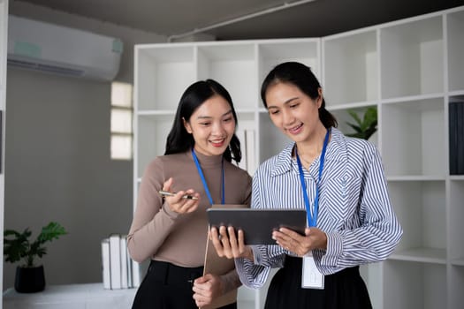 Two Asian businesswomen discussing work using a tablet in a modern office. Concept of teamwork and professional collaboration.