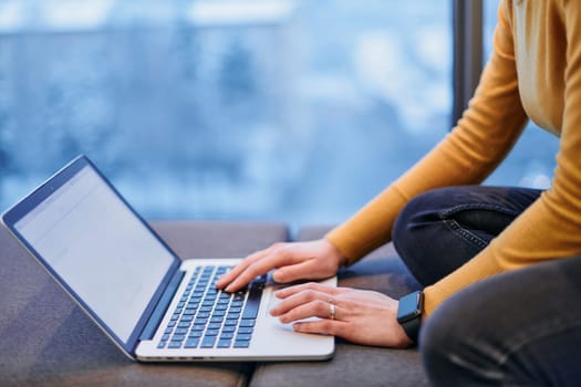 A businesswoman utilizes her laptop while seated by the window of a large corporate building, offering a picturesque view of the city skyline as her backdrop