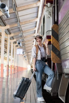 Happy young Asian female tourist carrying a backpack, holding a camera, preparing to wait for the train at the train station waiting for her vacation trip..