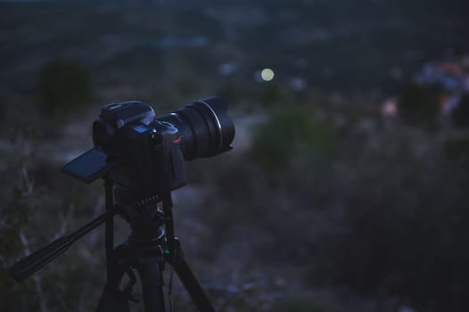 A black digital semi-mirror photographic equipment camera, with movable mockup touch screen, placed on tripod in nature, capturing steady picture or recording video of the city in the night and dawn.