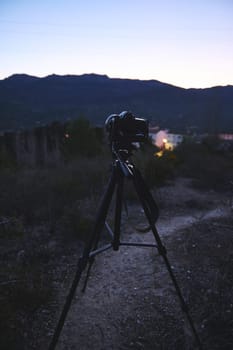 World photography day concept. Vertical shot of modern digital camera placed on tripod in nature outdoors, ready for capturing steady pictures or video of a village in mountains at dawn