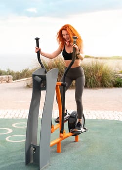 Fit woman working out on sport equipment of elliptical cross trainer on sunny day. Full body of strong redhead female athlete exercising at outdoor gym park. Healthy lifestyle.