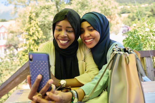 Two Middle Eastern Muslim women, adorned in hijabs, capture a moment of friendship and joy as they take selfies on a smartphone while seated in a natural setting.