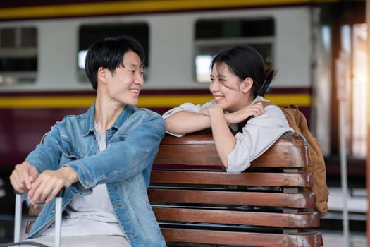 Happy young Asian couple carrying backpacks and cameras preparing to wait for the train at the train station waiting for their vacation trip together..