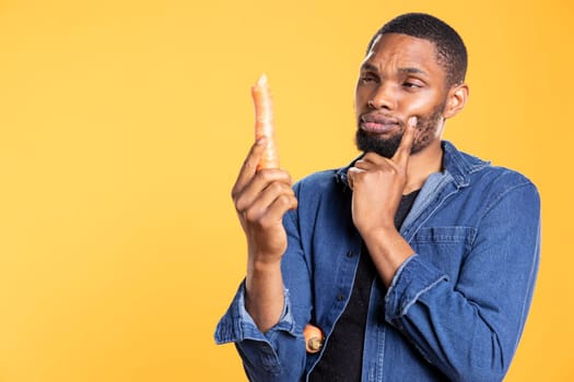 Pensive african american man thinking what recipe to cook with a carrot, holding freshly harvested vegetable for healthy eating concept. Confident person examining organic ripe veggies.