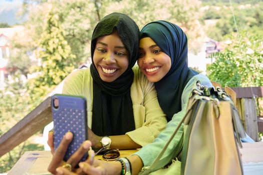 Two Middle Eastern Muslim women, adorned in hijabs, capture a moment of friendship and joy as they take selfies on a smartphone while seated in a natural setting.