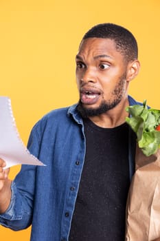 African american person is surprised of how many ingredients he needs for a special food recipe, shocked young adult in studio. Astounded male model shopping for organic produce.