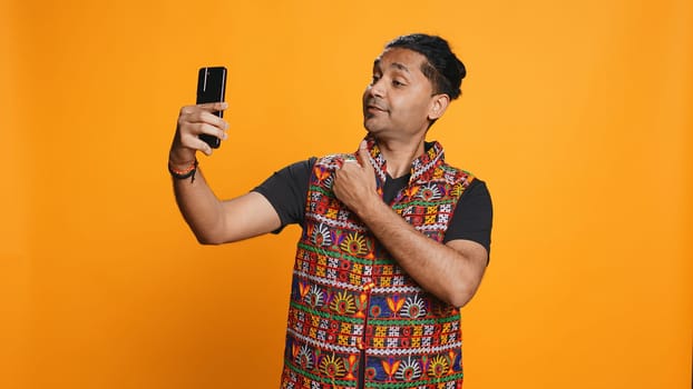 Indian narcissistic man using cellphone to take selfies from all angles. Vain social media user taking photos using phone selfie camera, smiling happily, studio background, camera B