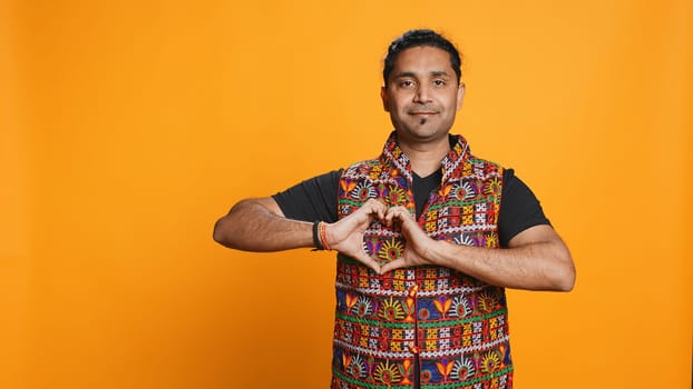 Portrait of jolly friendly indian man doing heart symbol shape gesture with hands. Cheerful nurturing person showing love gesturing, isolated over studio background, camera B