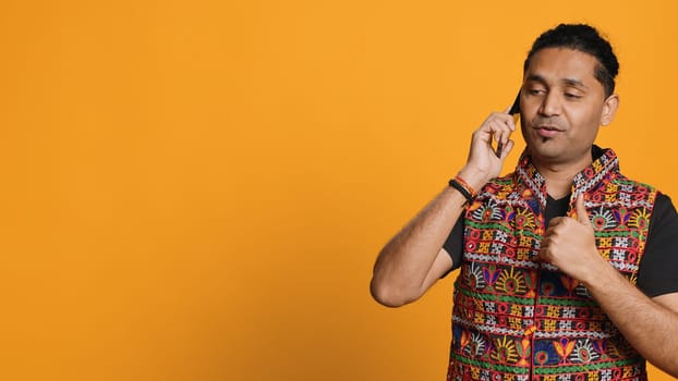 Happy man in traditional clothing chatting with best friend over telephone call. Upbeat person with colorful attire communicating with mate using phone, studio background, camera B