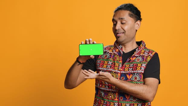 Content creator presenting green screen mobile phone, isolated over studio background. Indian person reviewing copy space mockup smartphone device for internet channel, camera A