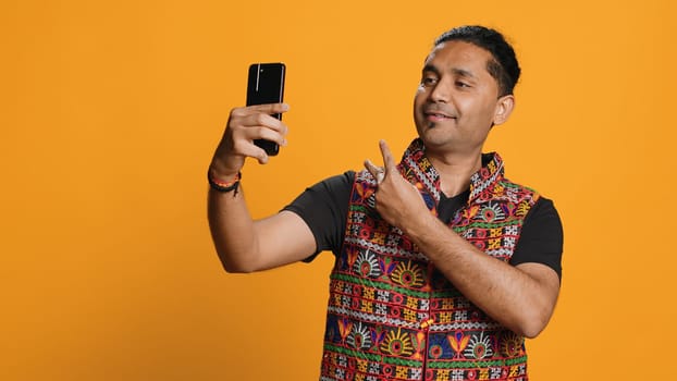 Indian narcissistic man using cellphone to take selfies from all angles. Vain social media user taking photos using phone selfie camera, smiling happily, studio background, camera A