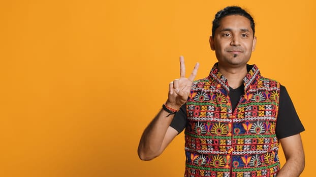 Portrait of upbeat man crossing fingers to make wish, hoping for good luck, isolated over studio background. Jolly indian person doing peace victory hand sign gesture, camera A