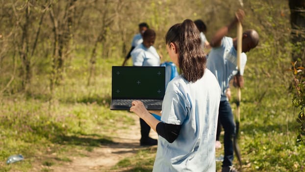 Environmental conservation volunteer holds a greenscreen laptop, working on cleaning up the forest with her team. Nature activists using isolated mockup and collecting rubbish. Camera A.