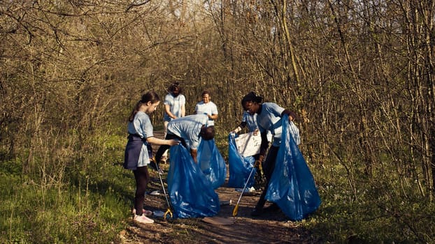 Group of volunteers working to clean the forest from garbage, collecting trash and plastic waste in bags. Environmental activists grabbing rubbish and recycling, support ecological justice. Camera B.
