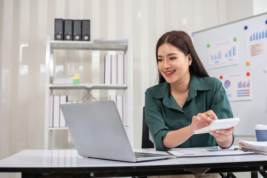 Asian businesswoman working on financial document with laptop on desk in office room.