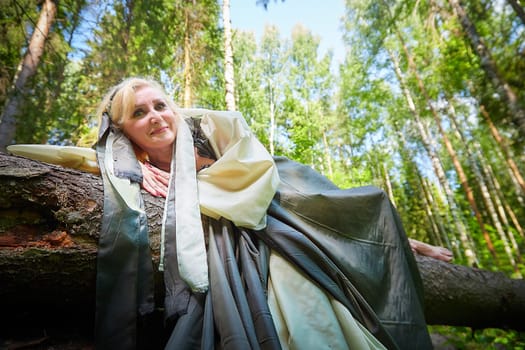 Adult mature woman 40-60 in a green long fairy dress in forest. Photo shoot in style of dryad and queen of nature. Fairy who loves nature and resting in summer big forest. Concept of caring for nature
