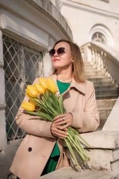 A woman wearing a tan coat and green dress holding a bouquet of yellow tulips. She is standing on a stone staircase
