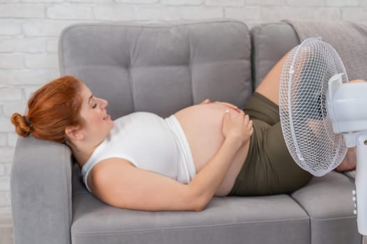 A pregnant woman is lying on the couch, stroking her tummy and enjoying the cool air from the electric fan