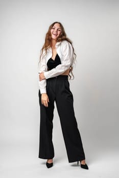 A young woman with long, wavy hair is caught in a moment of laughter, clad in a stylish white blouse, black trousers, and heels. She stands confidently with one hand on her hip against a neutral grey background, exuding happiness and a carefree spirit.