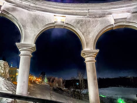 Winter Night View Through an Arched Stone Balcony Overlooking Lit Pathway. Arched balcony frames a snowy landscape under a night sky
