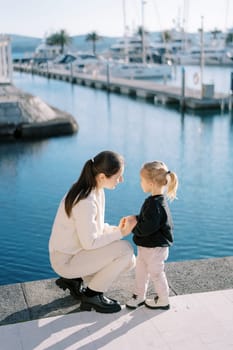 Mom holds the hands of a little girl squatting on the pier. High quality photo