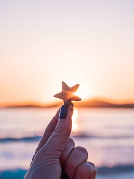 A hand with dark nail polish holds a starfish up towards the sunset, with the ocean and mountains in the background. The sky is filled with vibrant colors as the sun sets over the water.