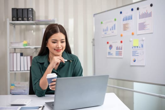 Asian businesswoman working on financial document with laptop on desk in office room.