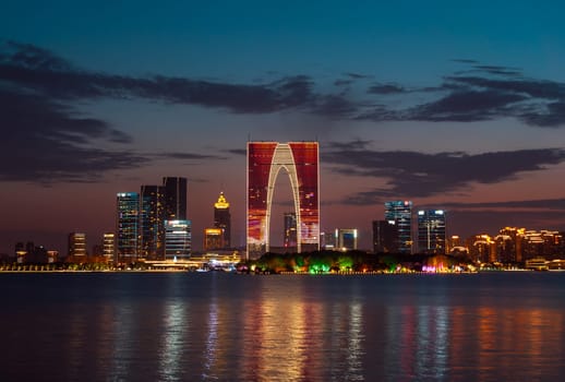 The Gate of the Orient, a distinctive skyscraper in Suzhou, China, is illuminated against the evening sky as it overlooks a calm lake. Surrounding buildings and city lights reflect on the water, creating a picturesque urban scene.