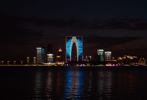 The Gate of the Orient, a distinctive skyscraper in Suzhou, China, is illuminated against the evening sky as it overlooks a calm lake. Surrounding buildings and city lights reflect on the water, creating a picturesque urban scene.