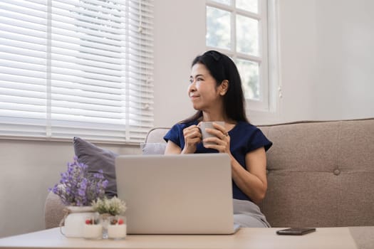 Happy Asian retired woman in her 60's enjoying social media on laptop while relaxing in living room..