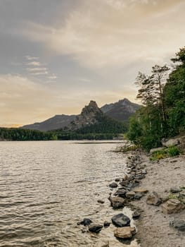 The warm glow of sunset illuminating the tranquil mountain lake, reflecting the rocky shoreline and lush green trees on the shore.