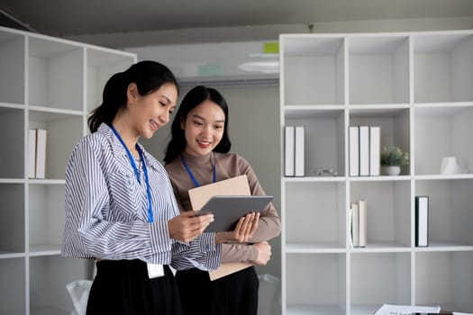 Two Asian businesswomen discussing work using a tablet in a modern office. Concept of teamwork and professional collaboration.