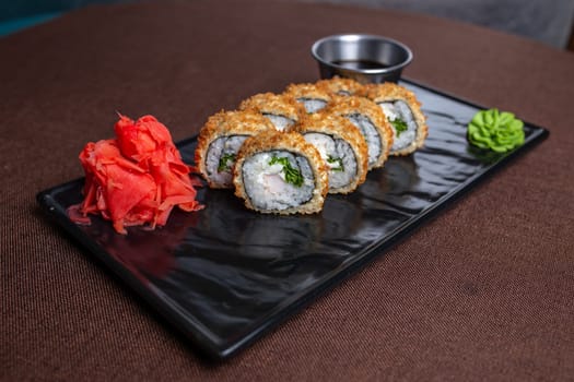 Deep-fried sushi roll with chopsticks and soy sauce on a black plate, made with rice, seaweed, fillings, and golden brown.