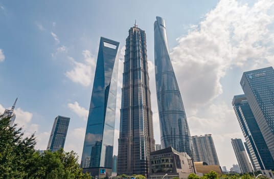 Three of Shanghais most iconic skyscrapers, including the Shanghai Tower, stand tall against a beautiful backdrop of clear blue skies and scattered white clouds.