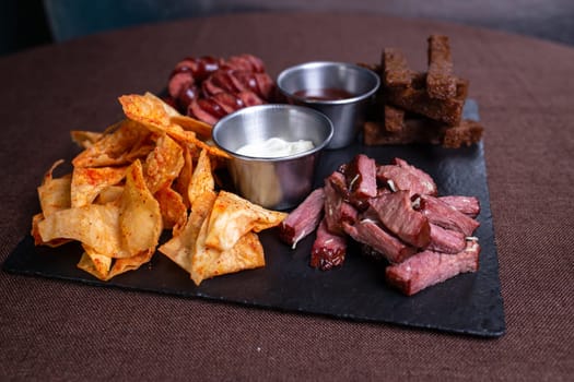 A top-down view of a tantalizing plate featuring bread, savory meat, delicious sausage, crispy chips, and flavorful dip.