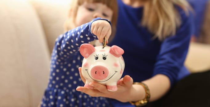 Child little girl arm putting pin money coins into happy pink faced piglet slot closeup. Making effective future needs savings collect dollar gift benefit present home leisure concept