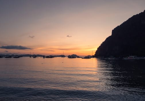 Multiple boats are anchored in a calm bay under a vibrant sunset. The sky glows with shades of orange and gold, while dark clouds create dramatic contrasts.