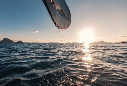 A kayak paddle drips water as the sun sets over a peaceful ocean, creating a tranquil and picturesque scene. The horizon is adorned with distant mountains, reflecting the golden hue of the setting sun.