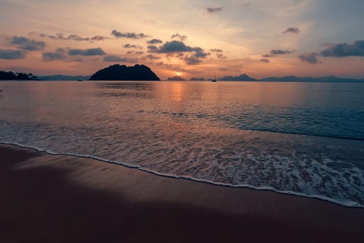 Golden and orange colors paint the sky as the sun sets over a serene beach. Gentle waves lap onto the shore, framed by distant islands and silhouettes of anchored boats.