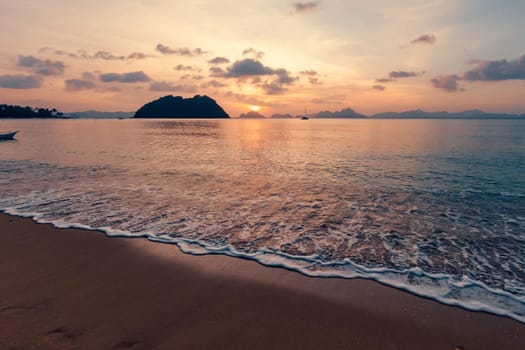 Golden and orange colors paint the sky as the sun sets over a serene beach. Gentle waves lap onto the shore, framed by distant islands and silhouettes of anchored boats.