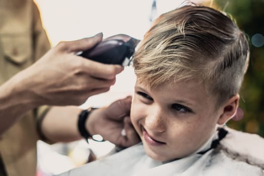 Barber shop blond boy customer. Hairdresser hand electric tool clipper. Child portrait real emotions.
