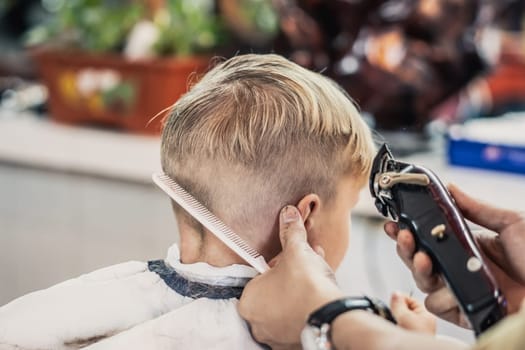 Barber shop blond boy customer. Hairdresser hand electric tool clipper comb. Back side view.