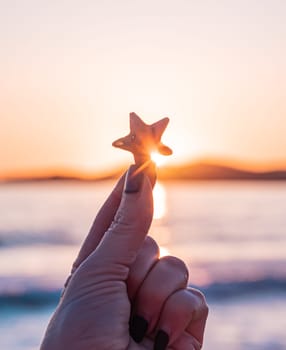 A hand with dark nail polish holds a starfish up towards the sunset, with the ocean and mountains in the background. The sky is filled with vibrant colors as the sun sets over the water.