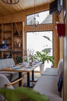 A rustic eatery with wood paneling, large windows, snowy landscape view, tables, chairs, fireplace, and bar, creating a warm ambiance.