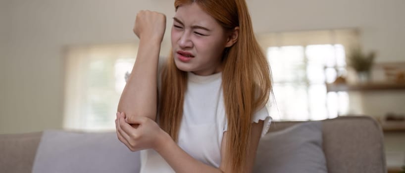 Young Asian woman experiencing severe arm pain while sitting on a sofa at home. Concept of health issues and discomfort.