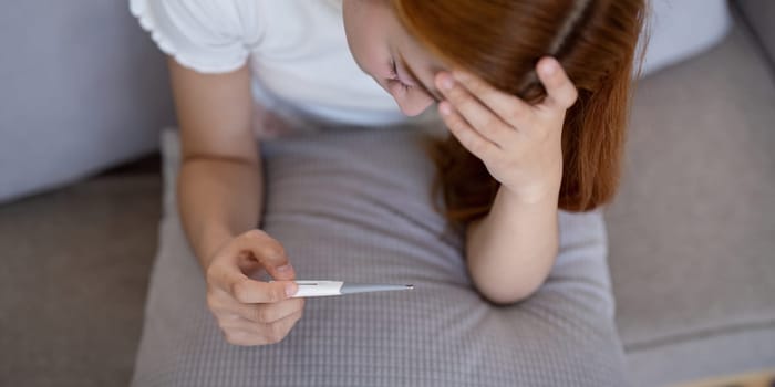 Young Asian woman with fever checking her temperature with a thermometer at home. Concept of illness and healthcare.