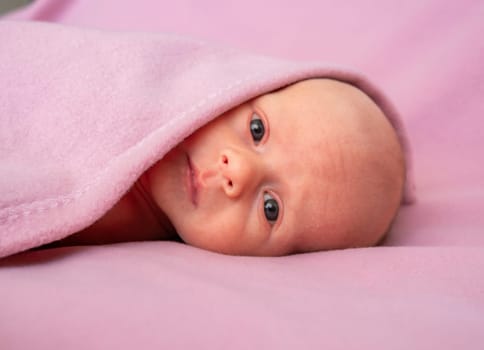 A newborn baby is comfortably wrapped in a soft pink blanket, gazing curiously at the surroundings.