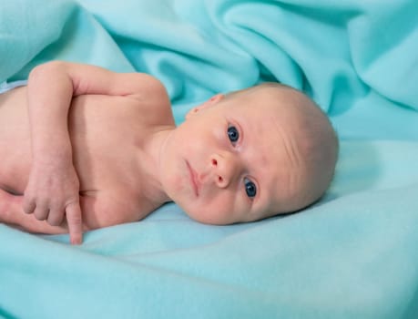 A newborn baby lies comfortably on a soft, aqua-colored blanket in a hospital room during the day. The baby gazes curiously at the camera, appearing calm and peaceful.